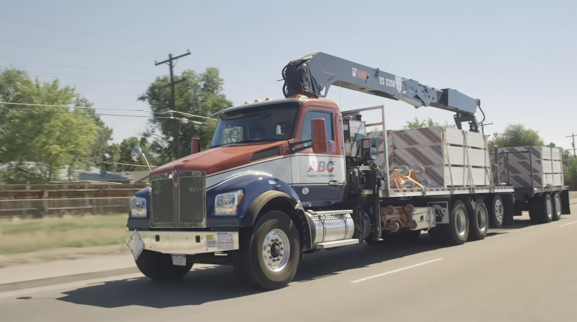 Hero Image, ABC Supply Interiors flatbed truck with crane hauling building materials along a road in daylight.